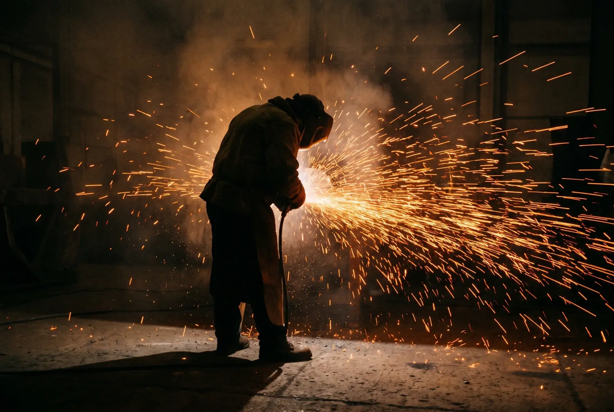 A welder silhouetted against a wall of arcing sparks in a dark warehouse.