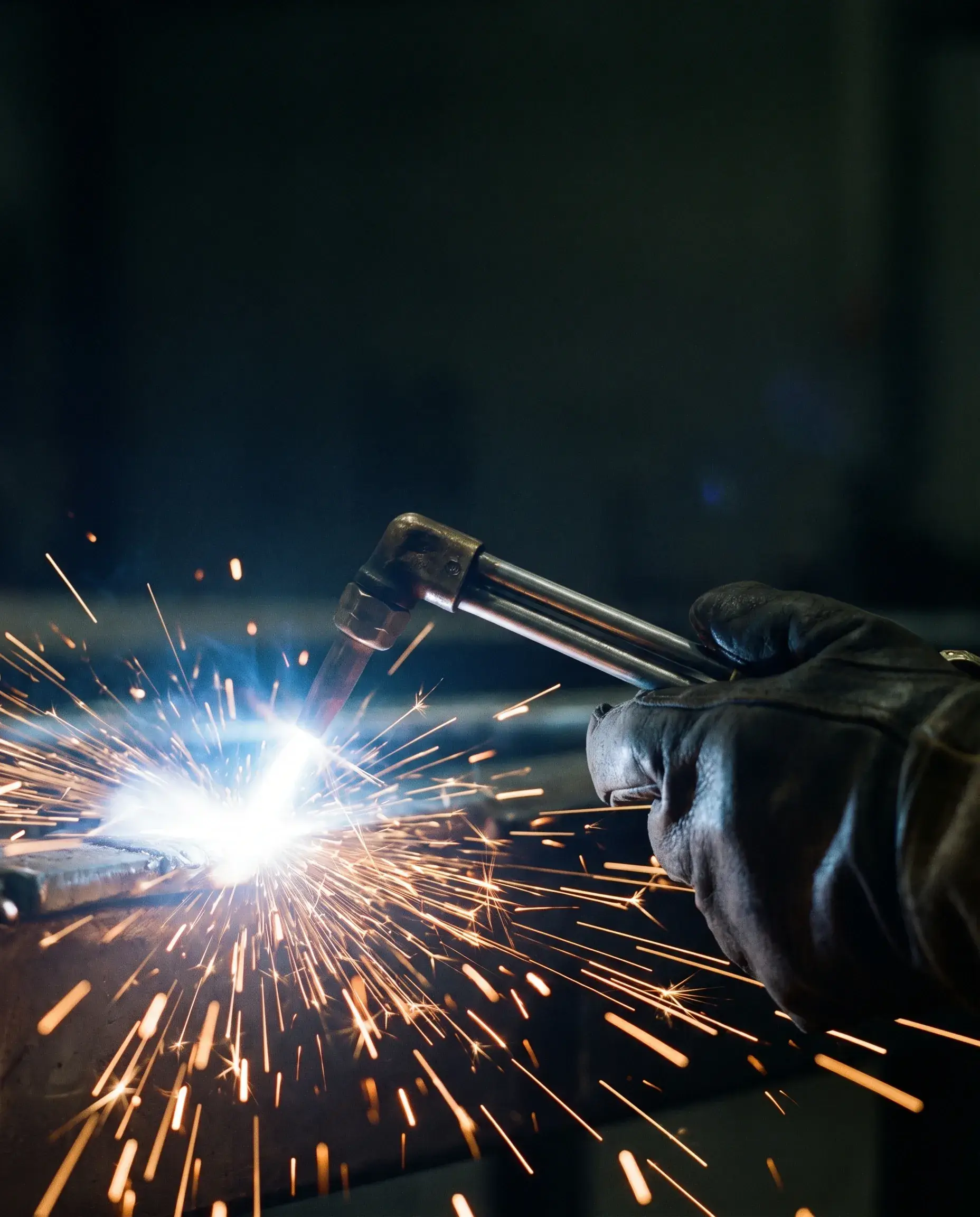 A gloved hand steadying a lit welding torch, sparks caught mid-flight against a dark workshop.