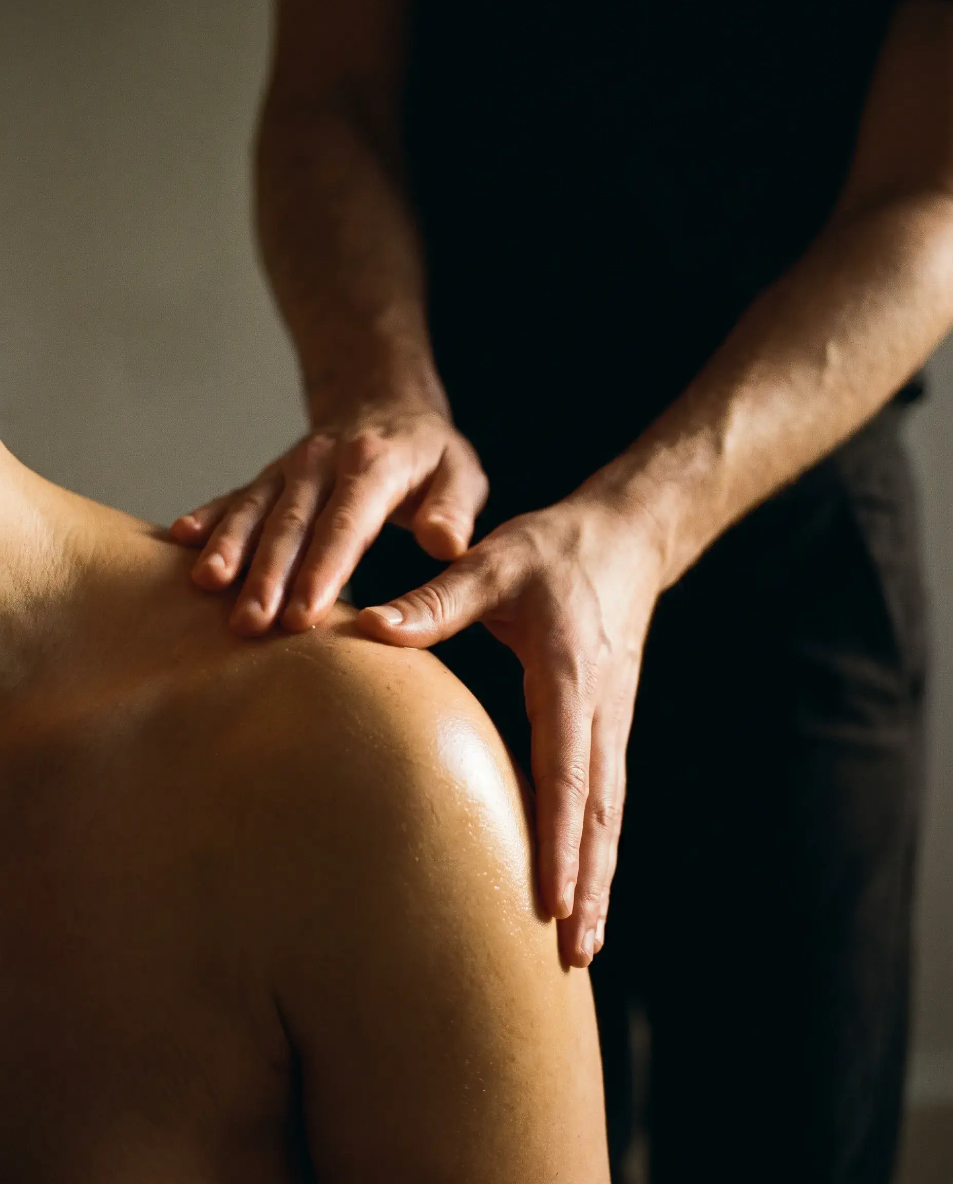 The hands of a massage therapist working a client’s shoulder under warm side light.